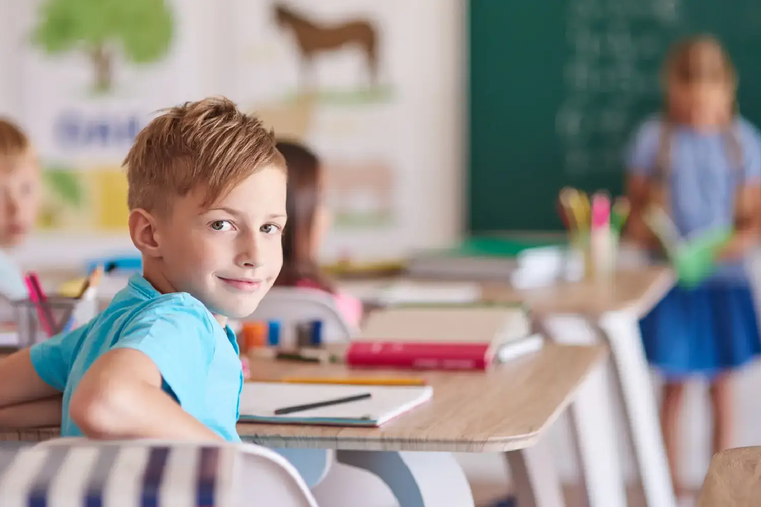 Aluno loiro, de camiseta azul, sorrindo, sentado em sua classe da escola no primeiro dia de aula.